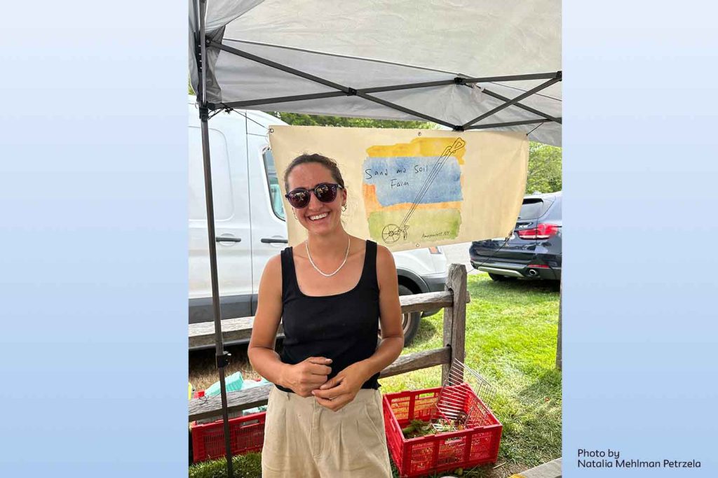 Andie Fortier in her farm's stand at a farmer's market. On the tent wall behind her is the farm's logo: a hand-painted plow on a watercolor wash of 3 colors.