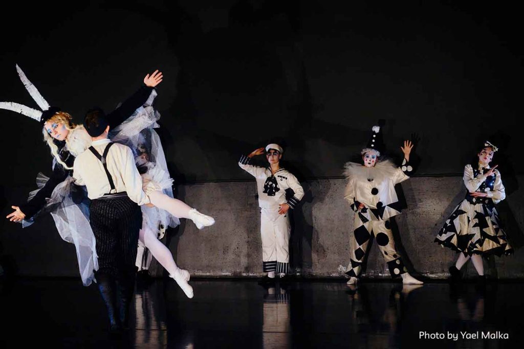 Production photo of Travesty Doll Play Ballez (after Copélia) in which four dancers dressed as marionettes hold poses as their characters in still mode (not wound up/playing). In the foreground, another dancer dressed in pants, shirt, and suspenders, carries one of the frozen marionettes back to the others. 